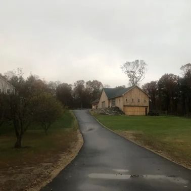 Asphalt driveway leads to a light-colored barn and green lawn on an overcast day. Trees line the right and left sides.