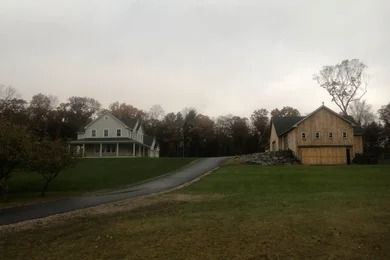 A two-story white house and wooden barn sit on a grassy hill, with a long driveway in front, under a gray sky.