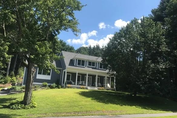 Two-story gray house with white trim, porch, and a grassy yard under a blue sky, partially obscured by trees.