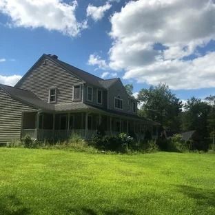 Two-story gray house with porch on a sunny, grassy lawn under a partly cloudy blue sky.