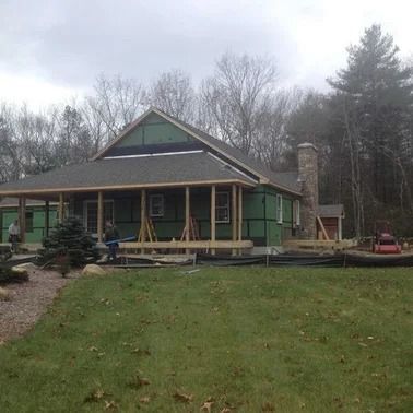 House under construction with green siding, wooden supports, chimney, and workers outdoors.