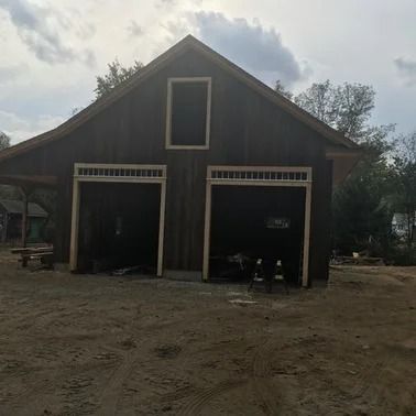 Dark brown barn with two garage doors and a small window under a cloudy sky.