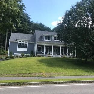 Blue house with a porch and green lawn, surrounded by trees under a blue sky.