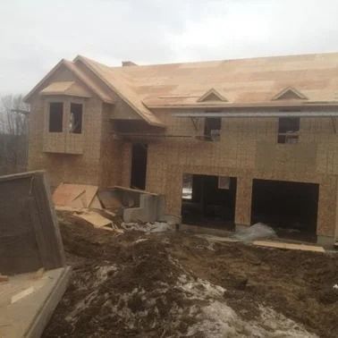 A two-story house under construction, framed with wood, has a partially built roof, garage doors, and windows.
