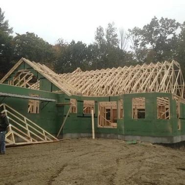 Construction site: wooden frame of a house with unfinished roof, surrounded by dirt and trees.
