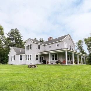 Gray two-story farmhouse with wrap-around porch on a large green lawn, under a cloudy sky.