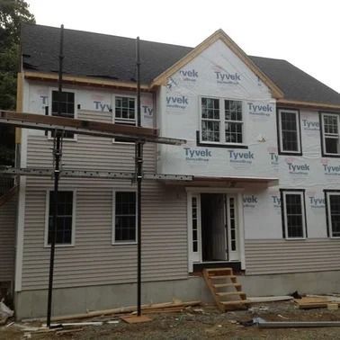 House under construction with gray siding, white windows, and black roof. Scaffolding is visible.