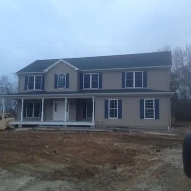 Two-story beige house with dark blue shutters and a porch, set in a construction site on an overcast day.