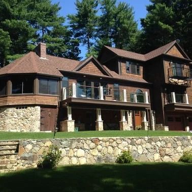 Brown wooden house with stone foundation, balcony, and lawn, surrounded by trees.