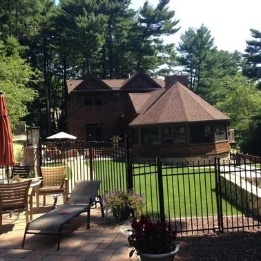 House with gazebo, black fence, and patio. Chairs and plants surround the yard with trees.