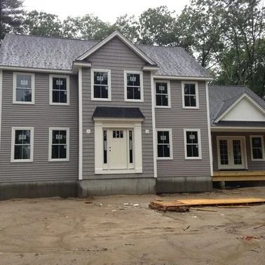 Gray two-story house with white trim. Windows on both floors, front door centered. Unfinished yard, wooded background.