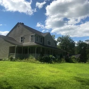 Two-story house with wrap-around porch, gray siding, green lawn under a partly cloudy blue sky.