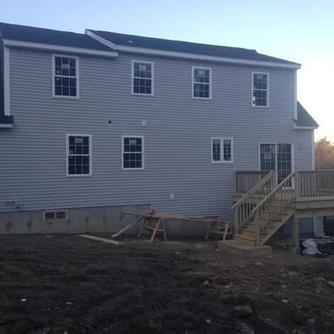 Back of a two-story gray house under construction with multiple windows, a deck, and exposed dirt.