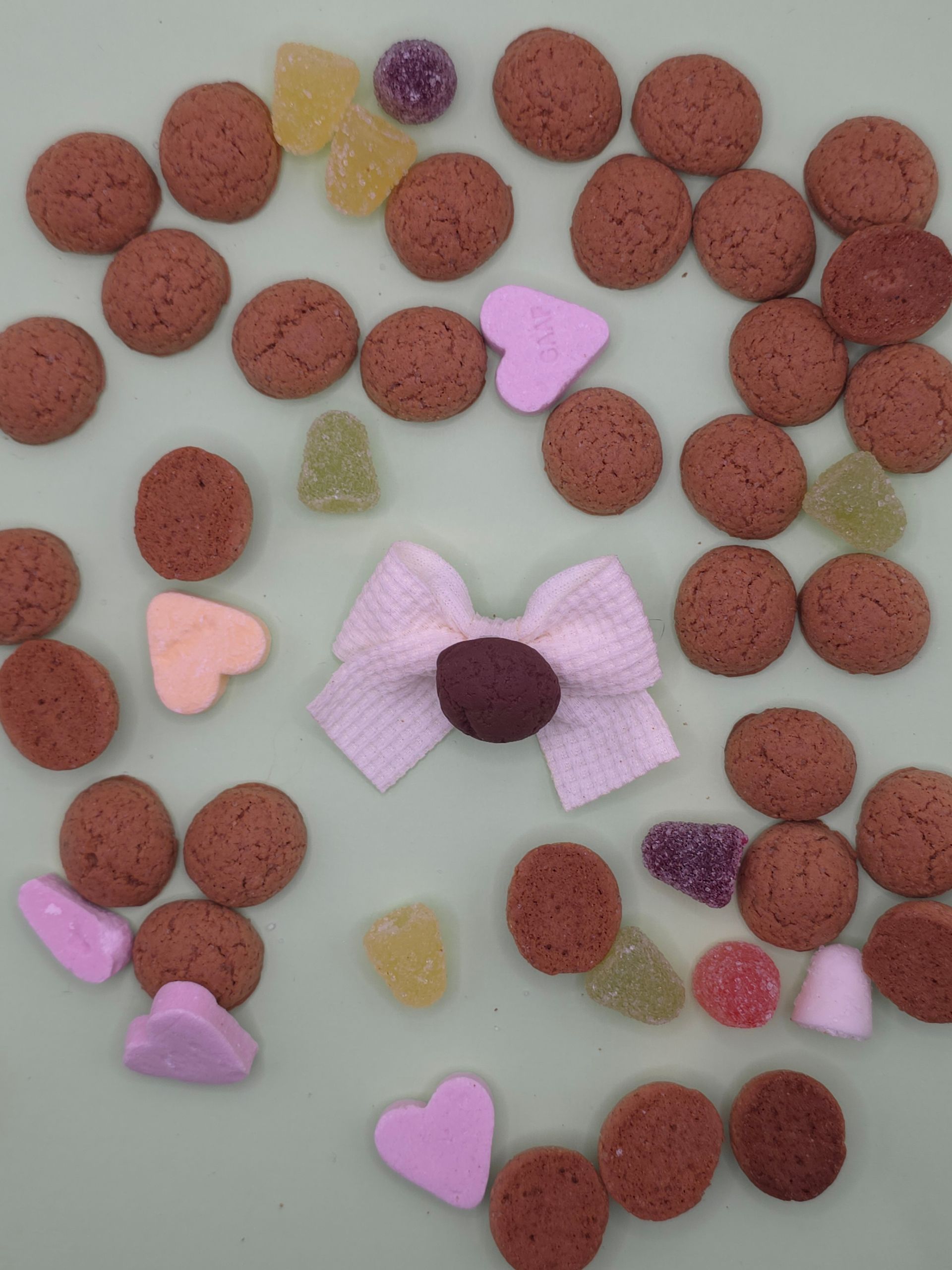 Verspreide rode cornflakesstukjes met pastelkleurige snoephartjes en een roze strikvormig snoepje op een witte ondergrond.
