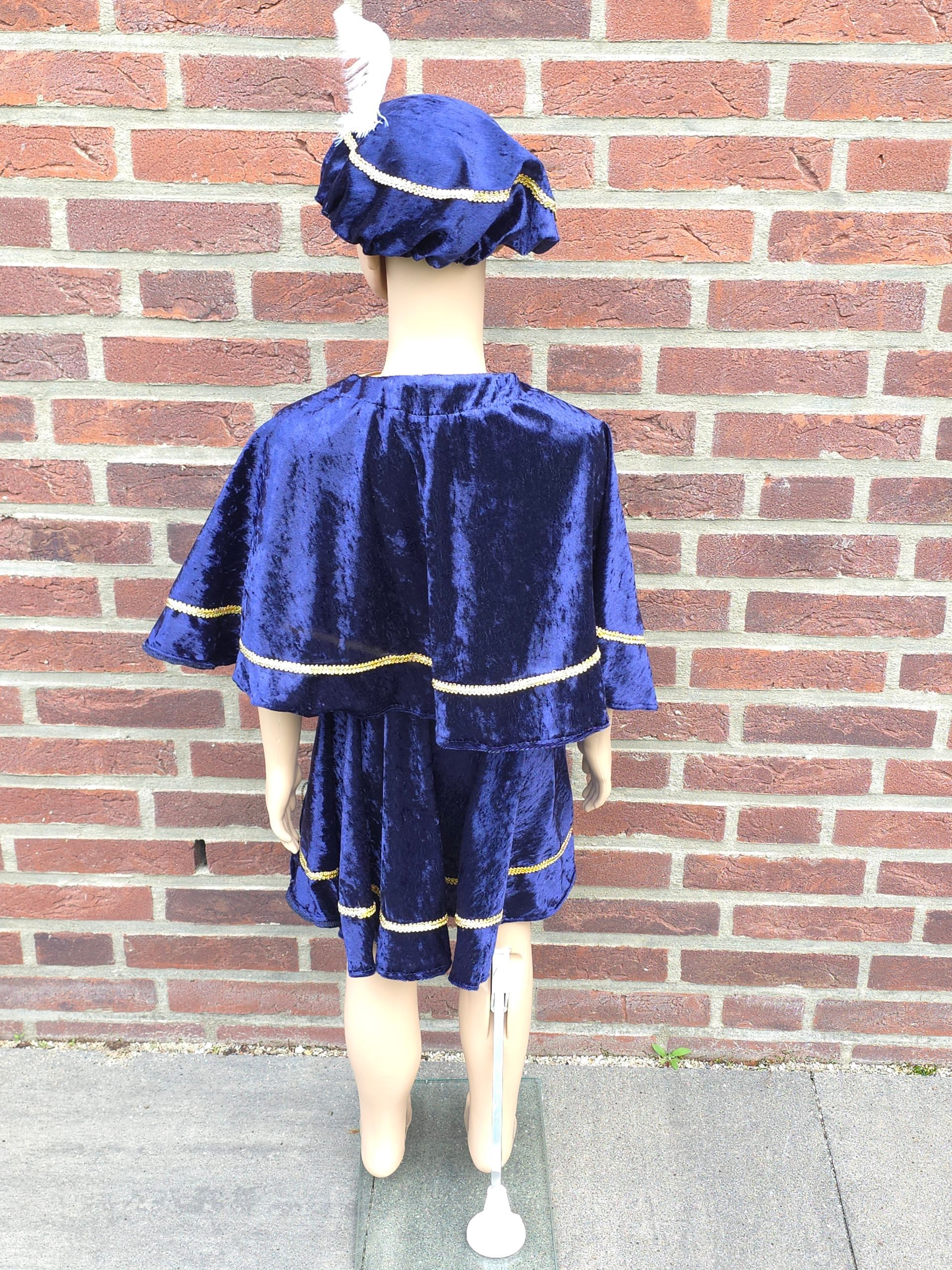 Een kind in een blauw fluwelen kostuum met gouden versiering en bijpassende hoed staat tegen een bakstenen muur.
