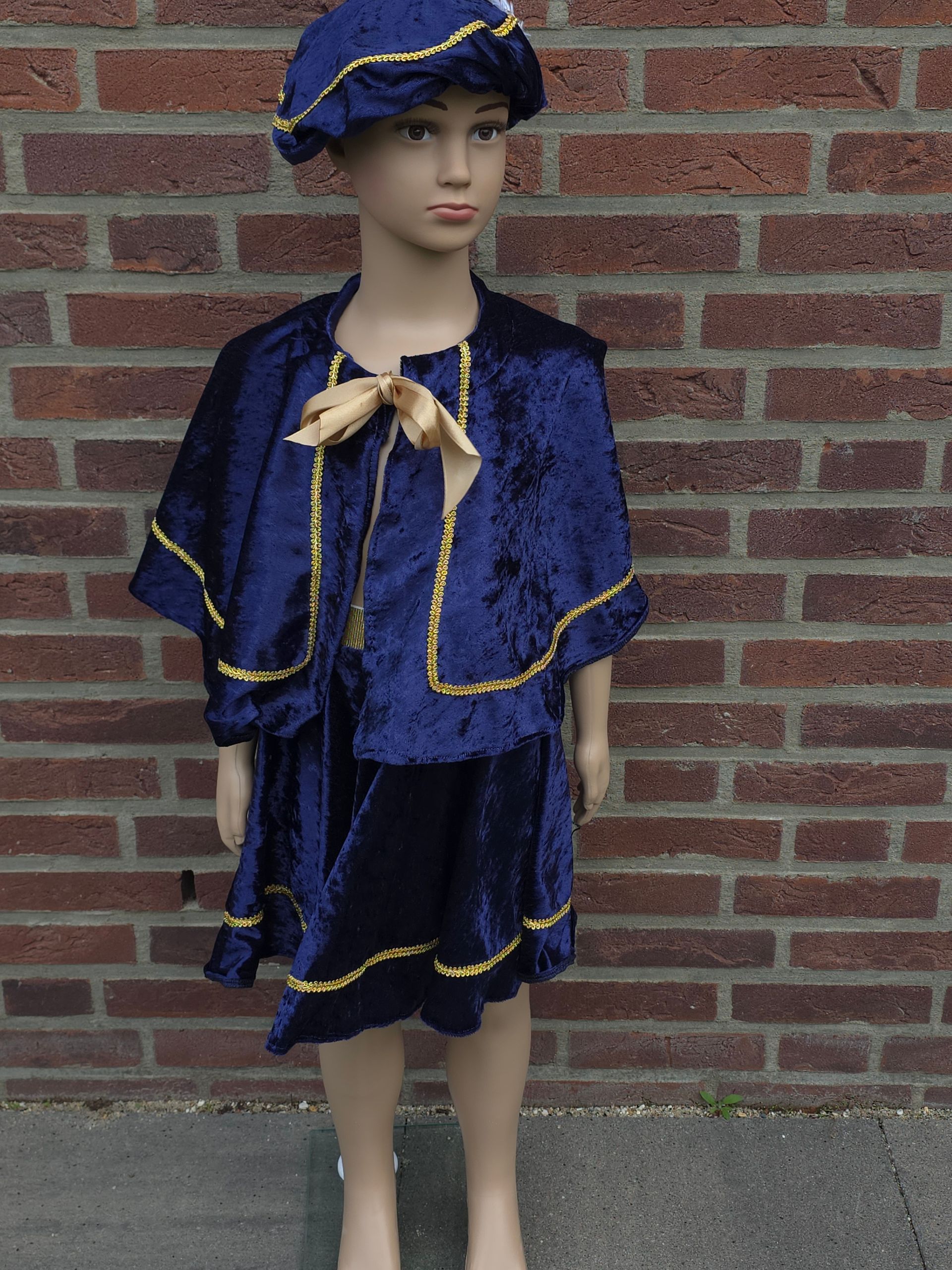 Een kind in een blauw fluwelen kostuum met gouden versiering en bijpassende hoed staat tegen een bakstenen muur.