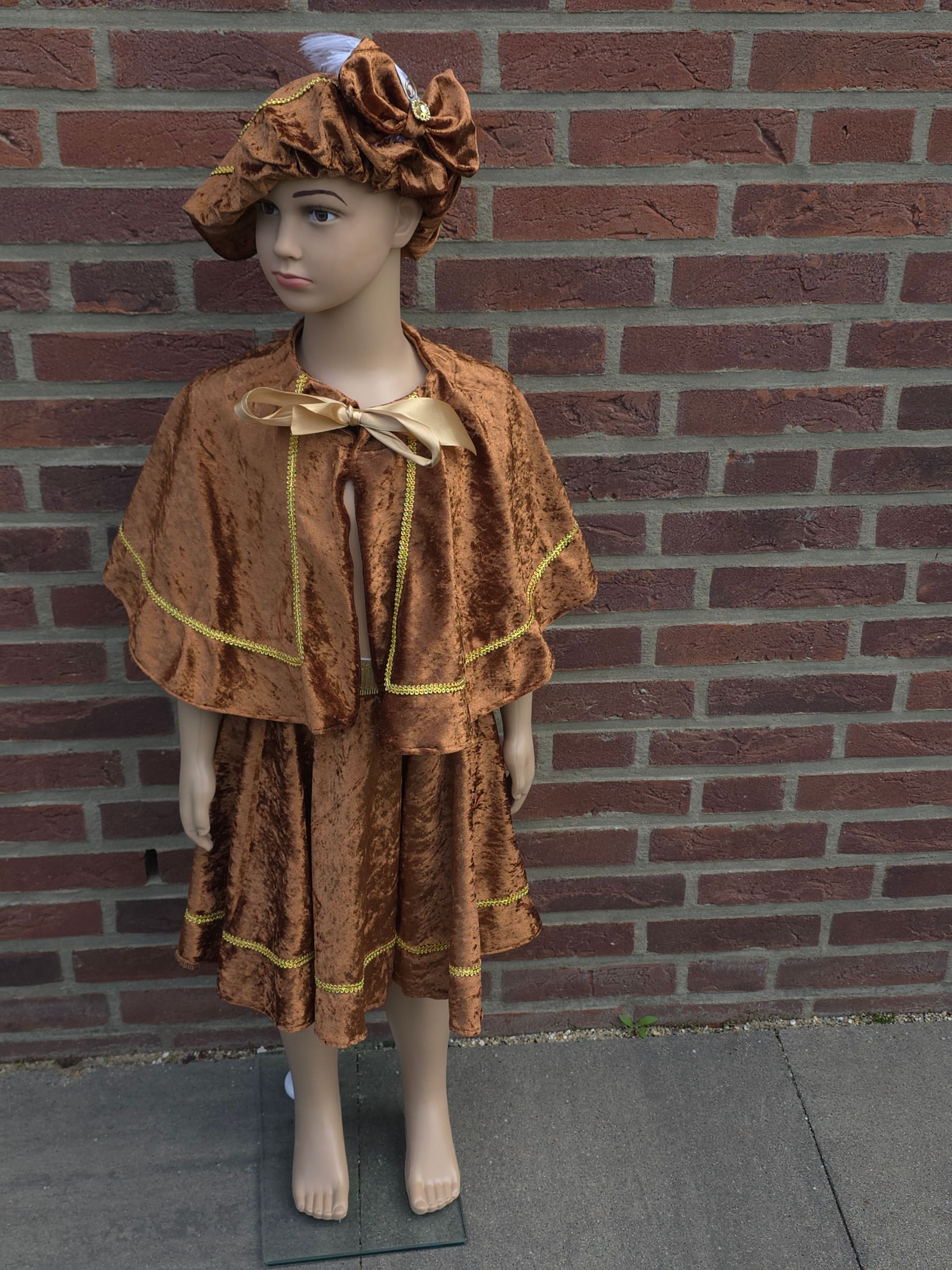 Een kindermannequin in een gouden jurk met een witte kanten kraag staat tegen een bakstenen muur.