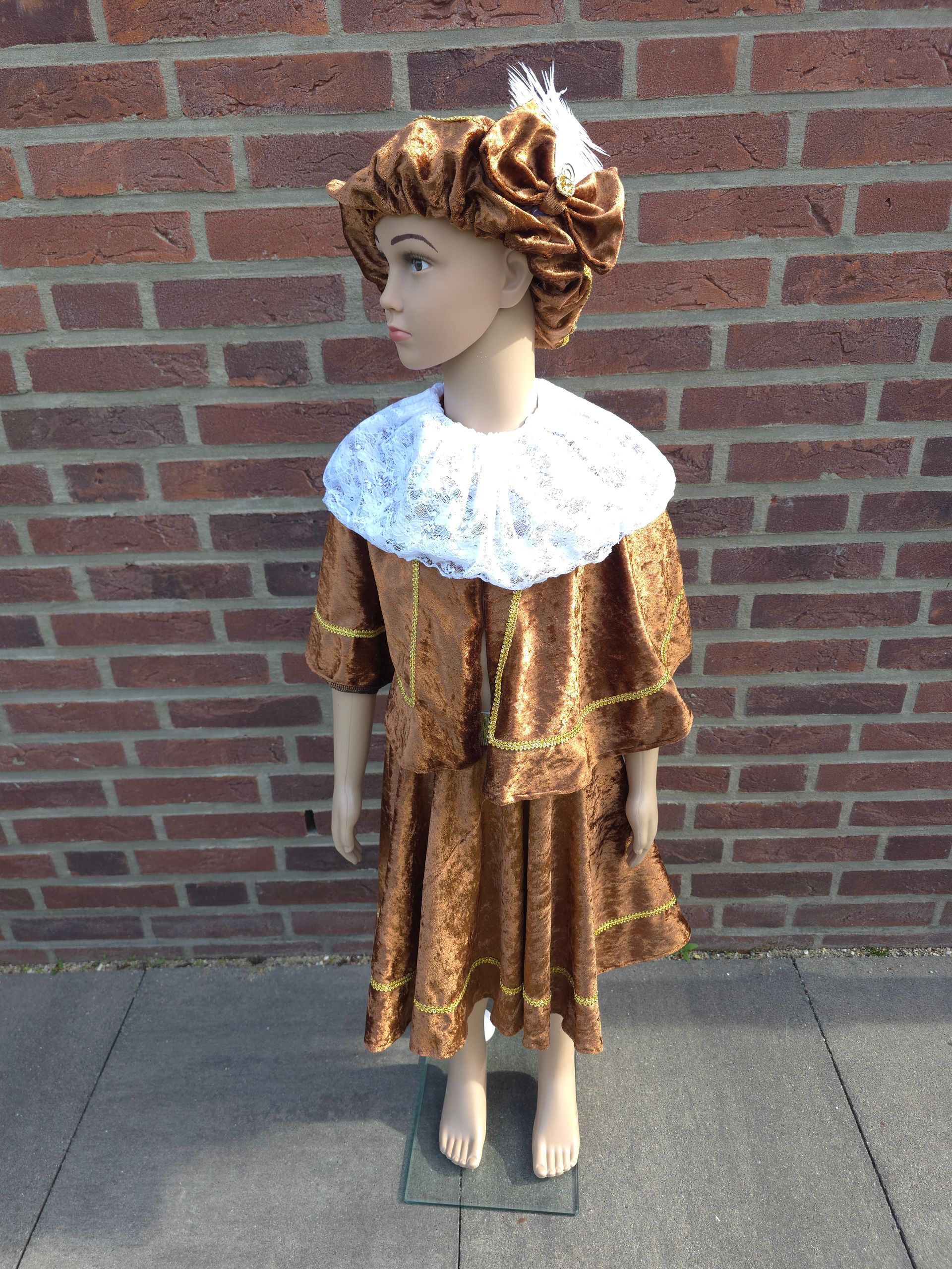 Een kindermannequin in een gouden jurk met een witte kanten kraag staat tegen een bakstenen muur.