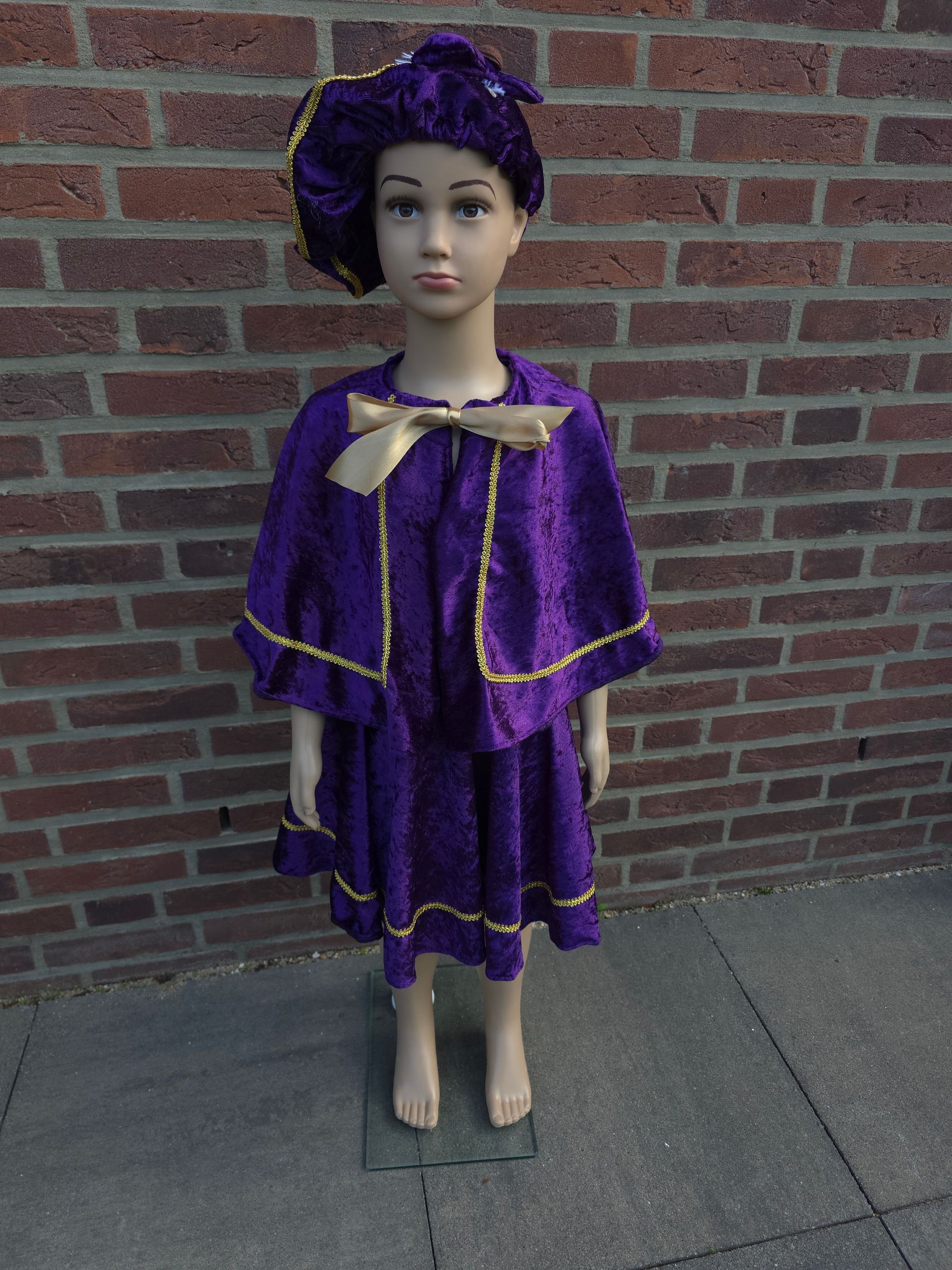 Mannequin in a purple dress and matching hat standing on a sidewalk against a brick wall