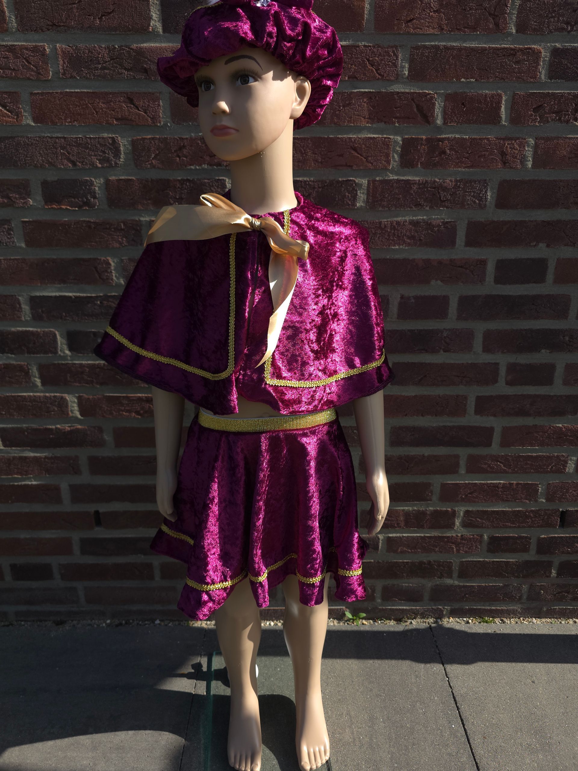 Een mannequin, gekleed in een glanzende paarse outfit en hoed, staat tegen een bakstenen muur in het zonlicht.