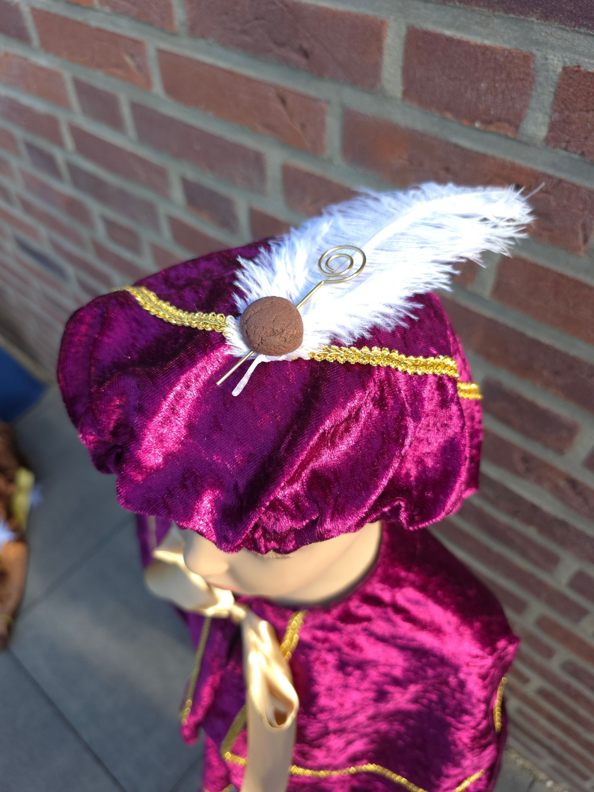 Paarse fluwelen verkleedhoed met witte veer en gouden versiering tegen een bakstenen muur