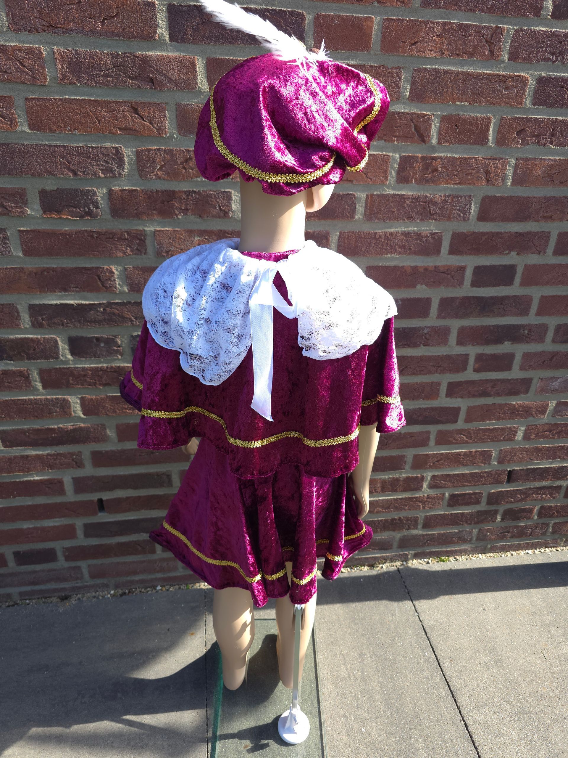Een mannequin, gekleed in een glanzende paarse outfit en hoed, staat tegen een bakstenen muur in het zonlicht.