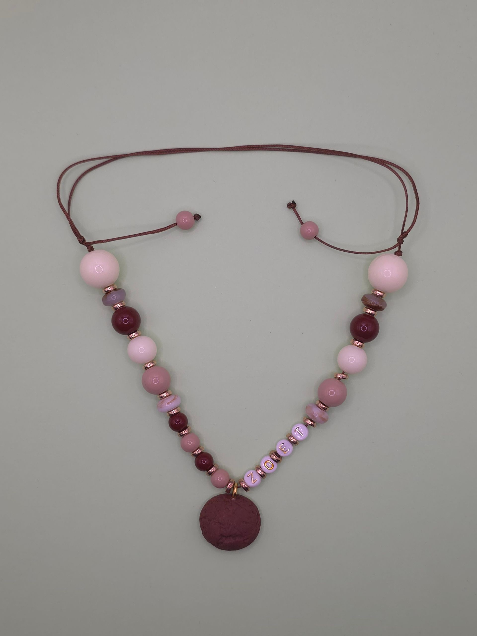 Roze kralenketting met letterkralen en een ronde bordeauxrode hanger op een grijze achtergrond.