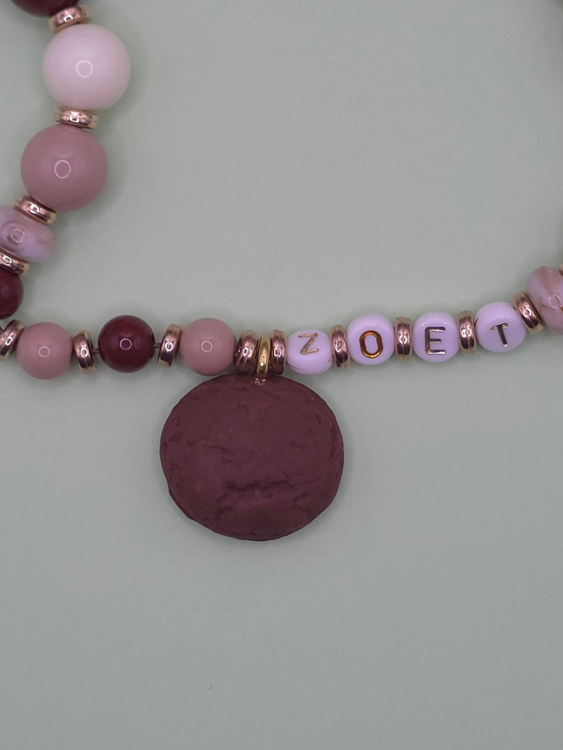Roze kralenketting met letterkralen en een ronde bordeauxrode hanger op een grijze achtergrond.
