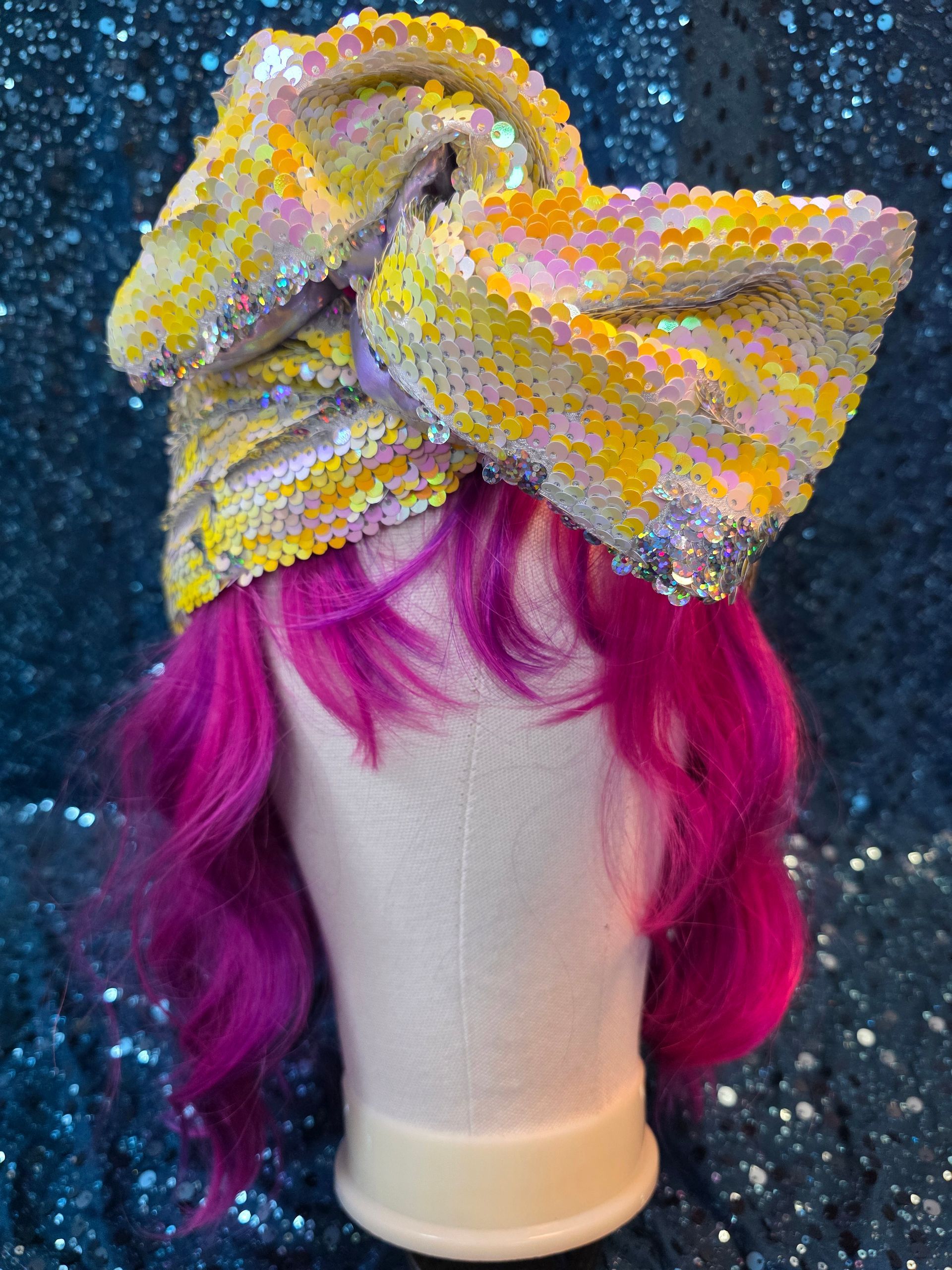 Wit hoofd met een felmagenta pruik en een glinsterende gele strik als hoofdband, tegen een donkere achtergrond vol glitters