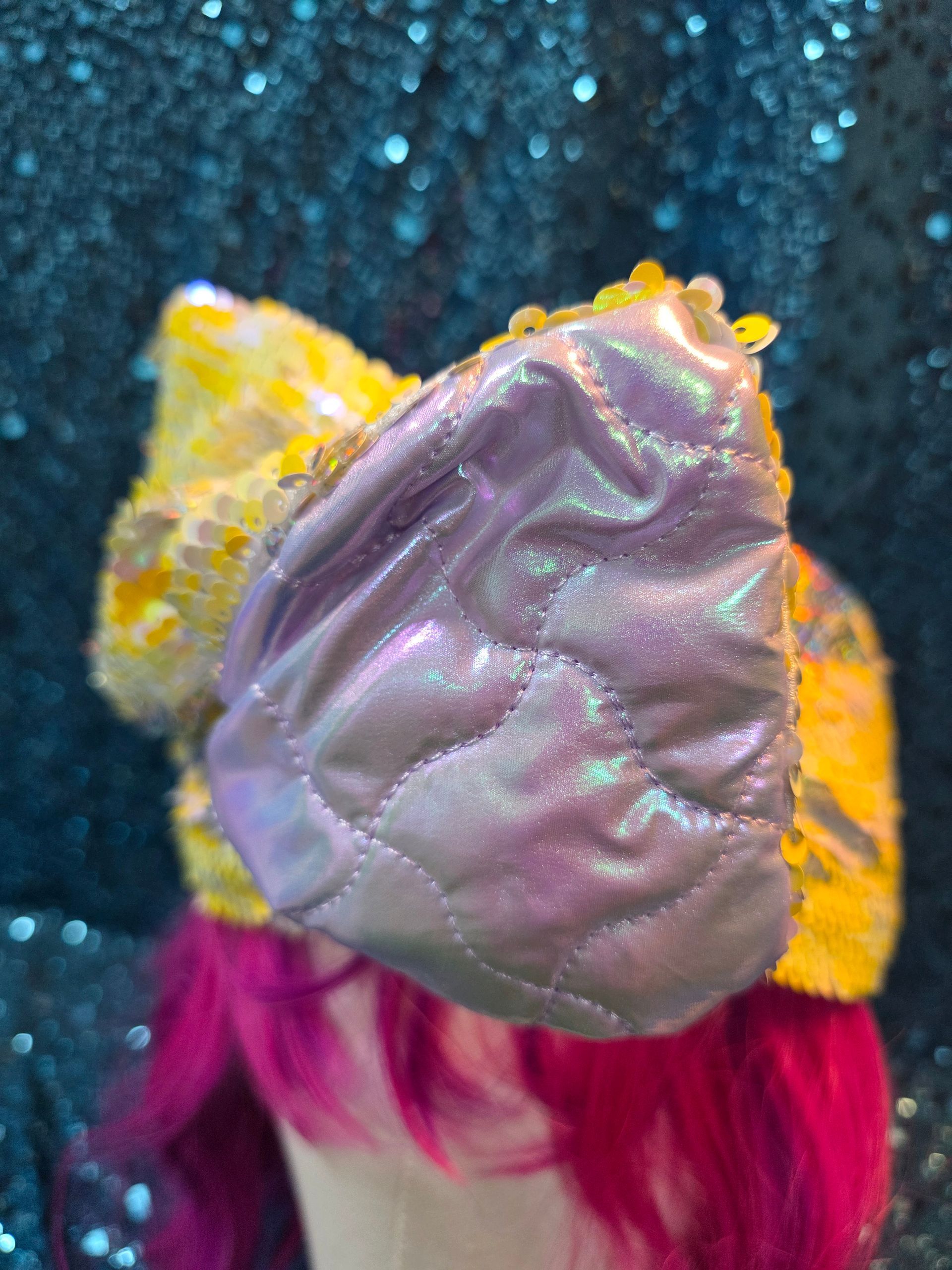 Wit hoofd met een felmagenta pruik en een glinsterende gele strik als hoofdband, tegen een donkere achtergrond vol glitters
