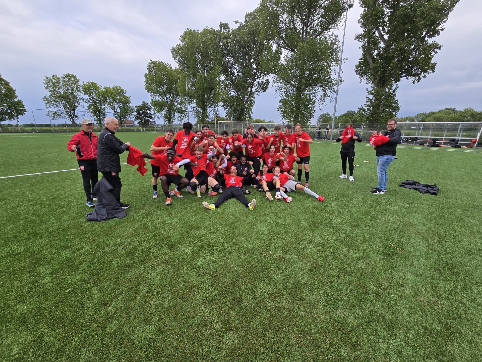 Een jeugdvoetbalteam in rode shirts poseert voor een groepsfoto op een grasveld met bomen op de achtergrond.