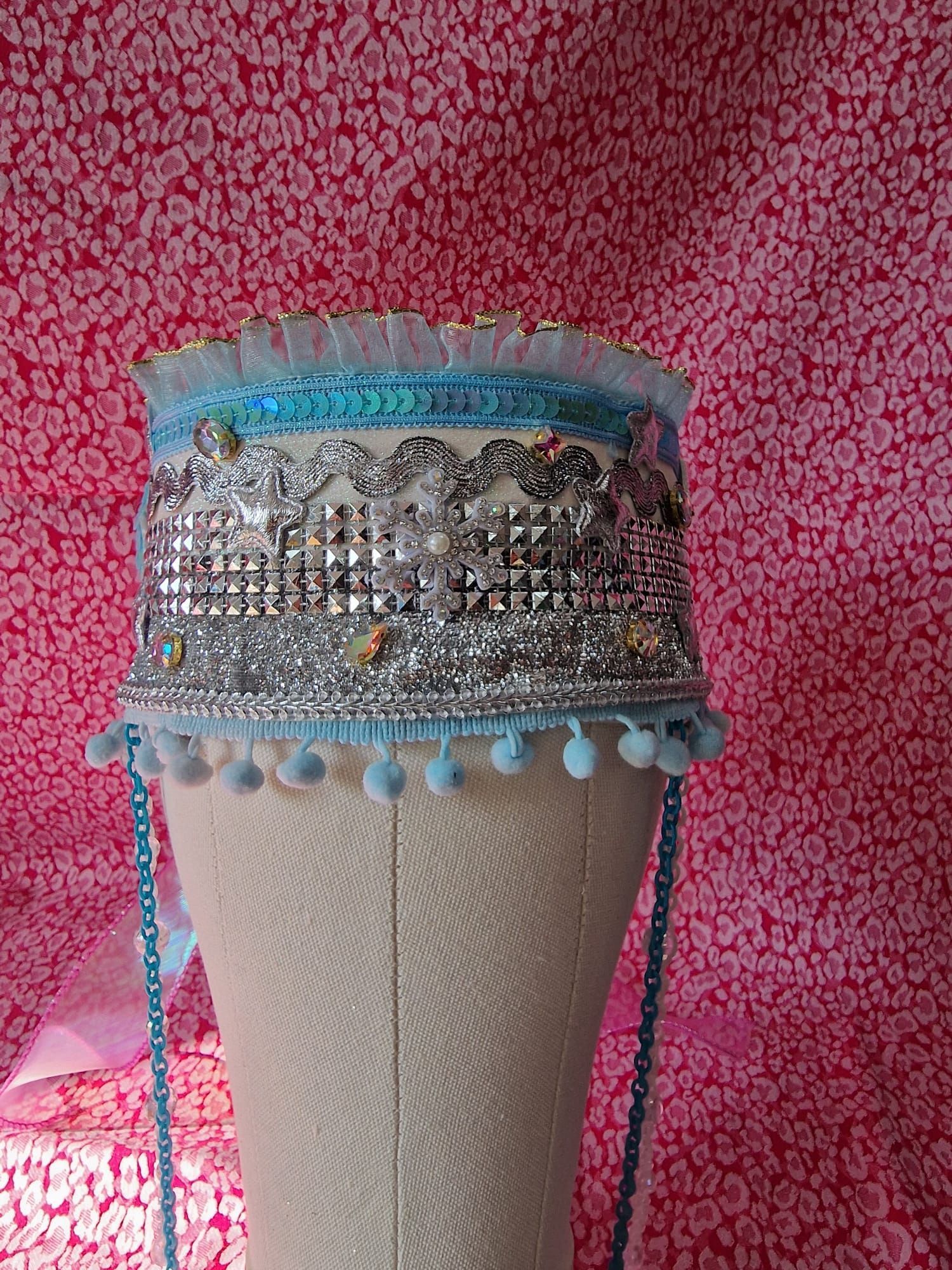 Decoratieve hoed met zilveren en turkooizen kralen op een paspop tegen een roze achtergrond met patroon.