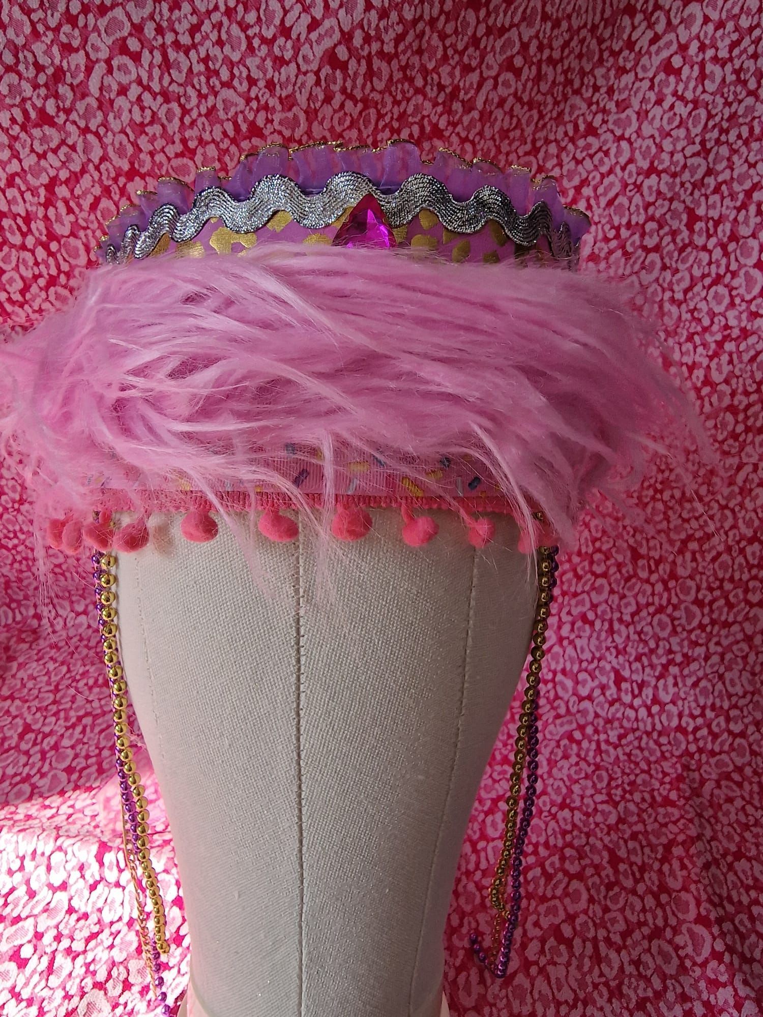 Roze hoed met veren en een met juwelen versierde band op een witte mannequin tegen een roze, gestructureerde achtergrond.