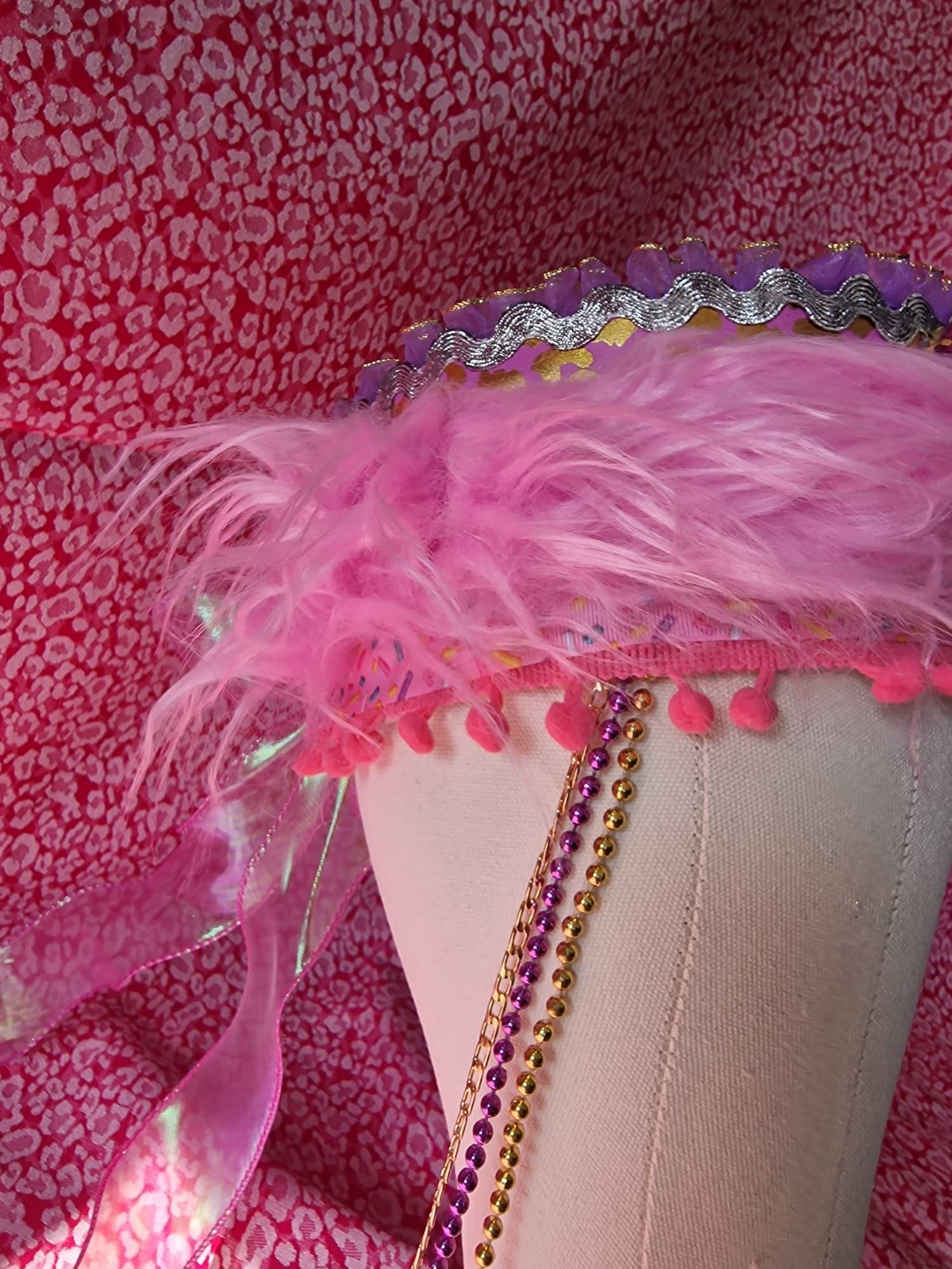 Roze hoed met veren en een met juwelen versierde band op een witte mannequin tegen een roze, gestructureerde achtergrond.