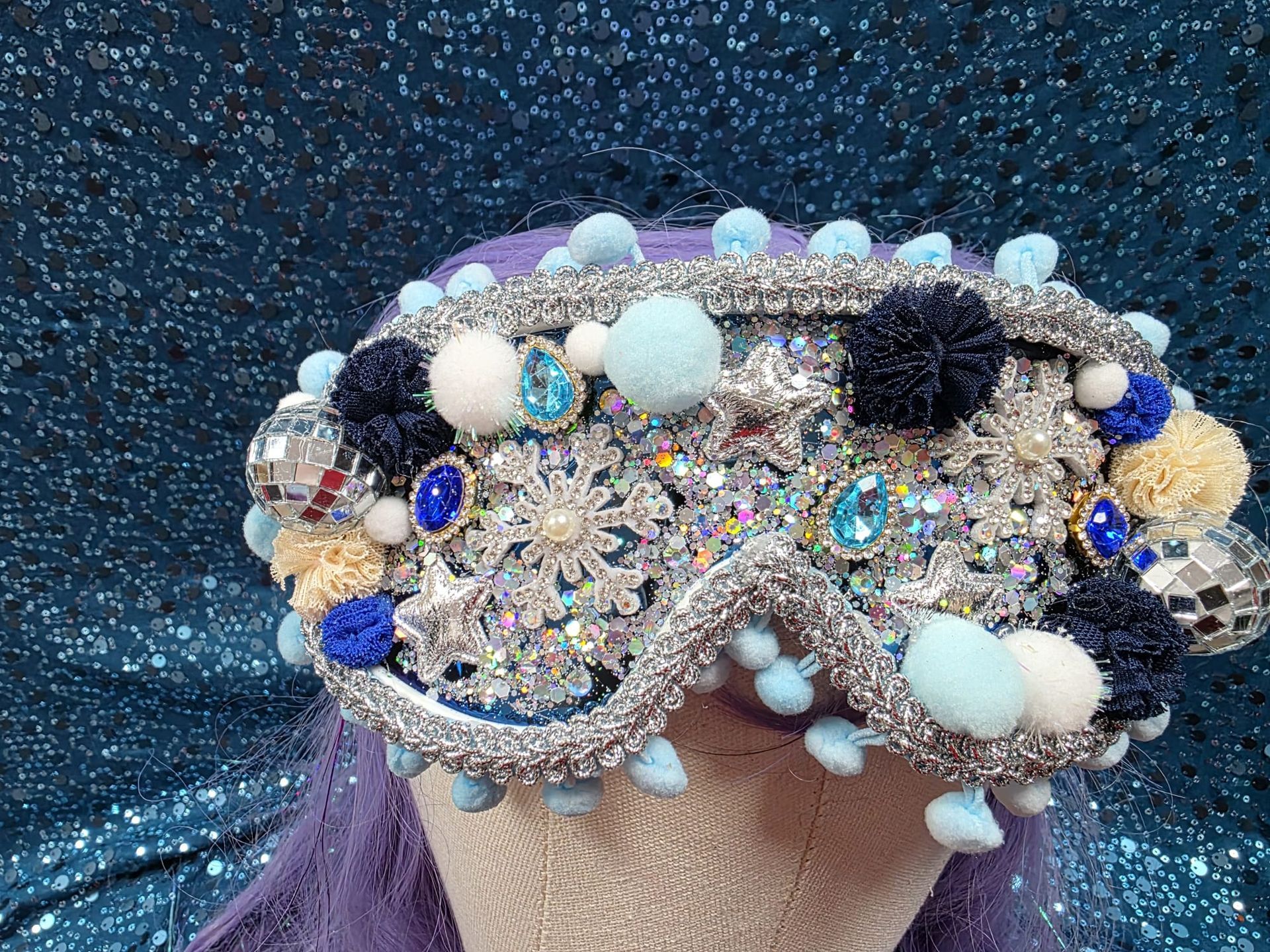 Een met kralen en juwelen versierde skibril met blauwe, witte en zwarte pompons op een glinsterende blauwe achtergrond.