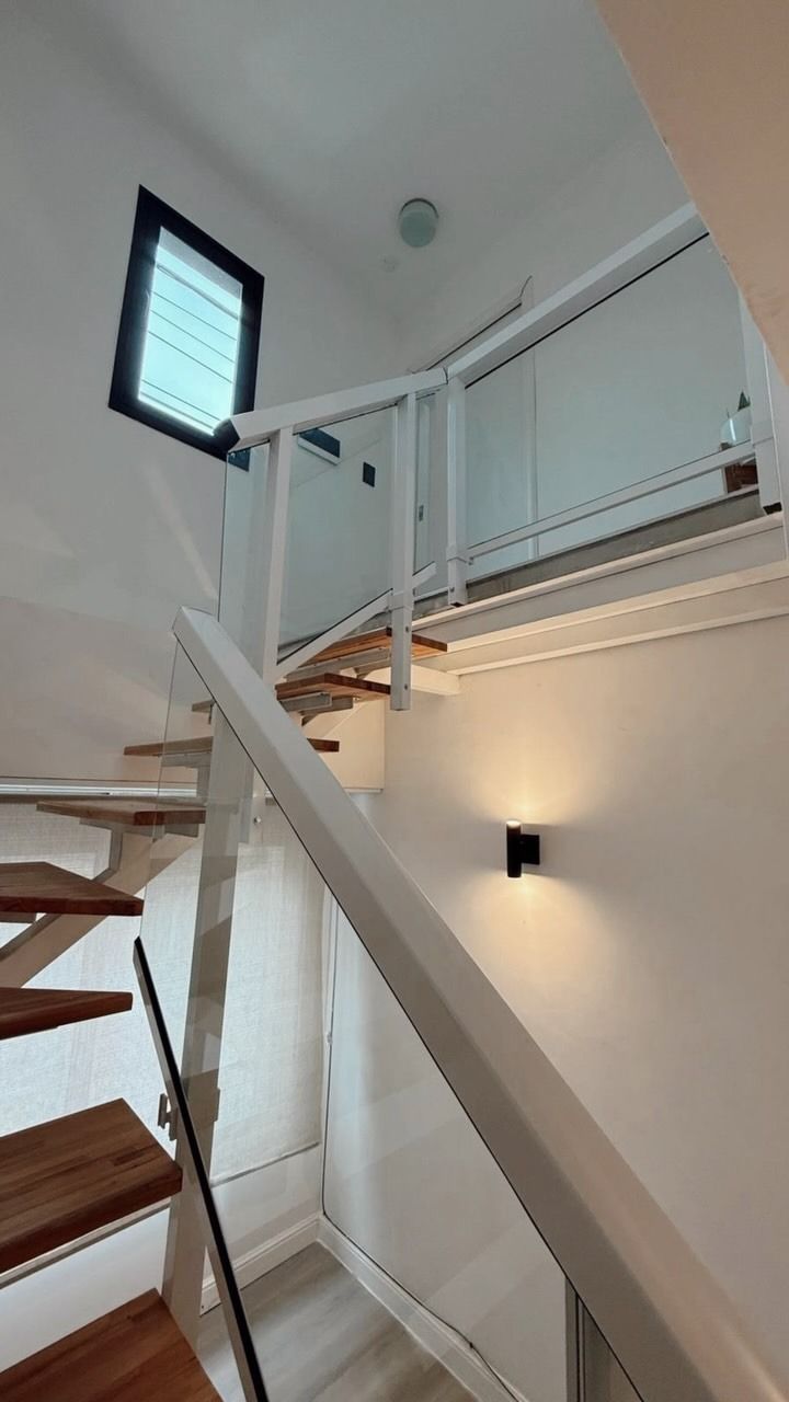 Rellano de escalera con barandilla blanca, escalones de madera, aplique de pared y una pequeña ventana en la parte superior.