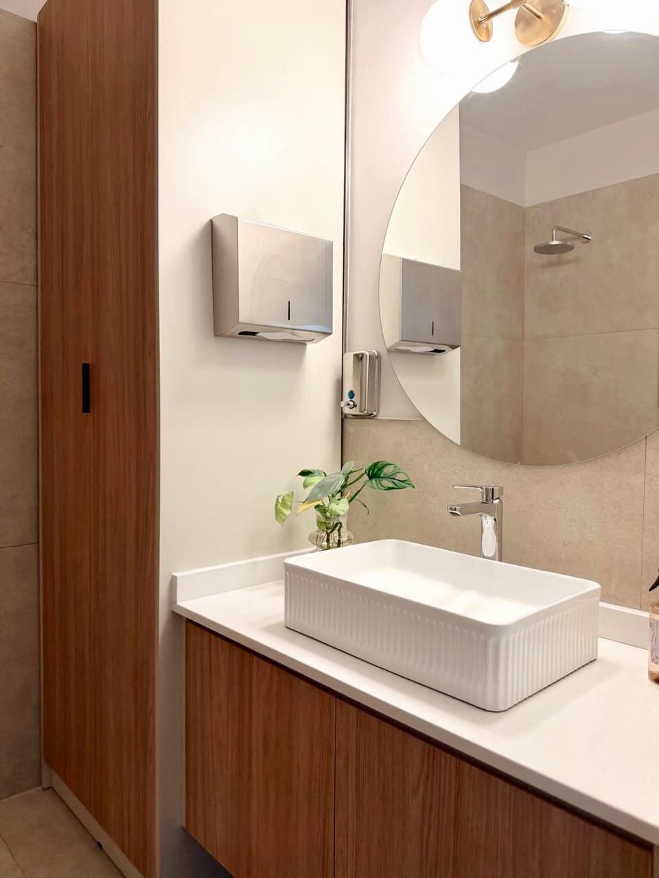 Tocador de baño moderno con gabinetes de madera, encimera blanca, lavabo sobre encimera, espejo y aplique de pared.