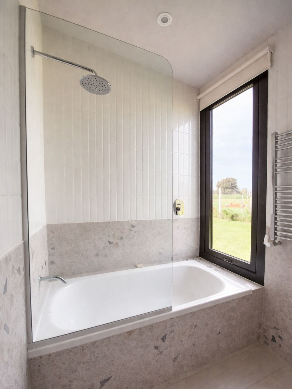 Baño moderno con bañera blanca, cabezal de ducha y gran ventanal con vistas a un campo de hierba.