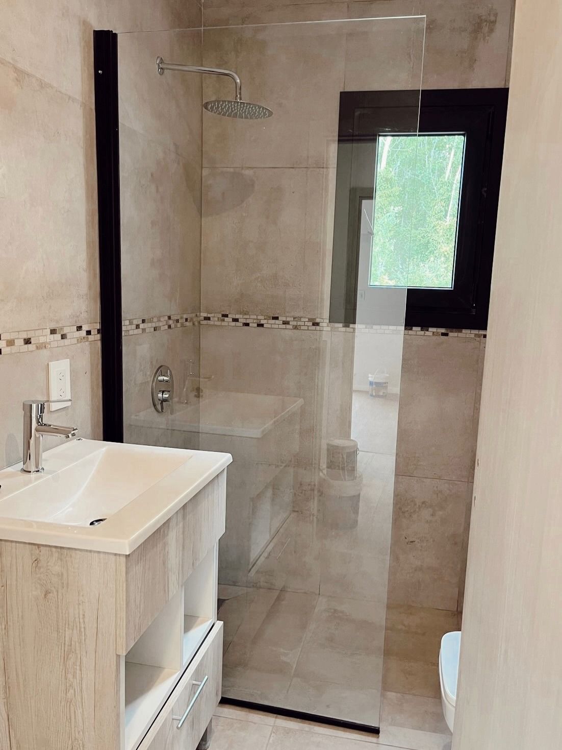 Baño moderno con ducha de cristal, paredes de azulejos claros y lavabo blanco.