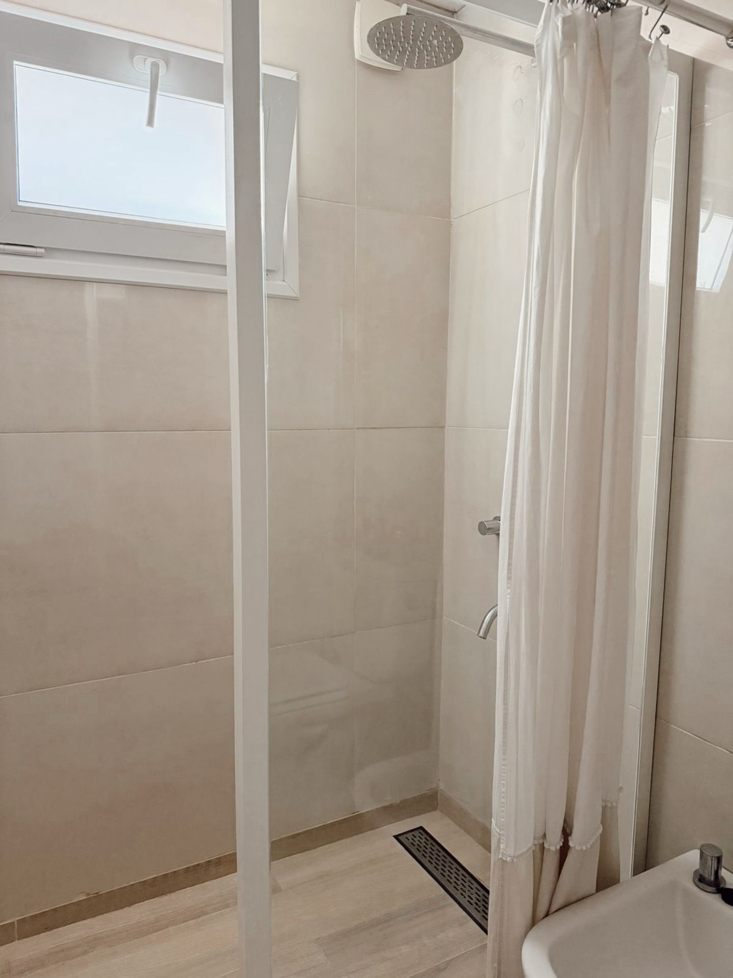 Pequeña ducha con azulejos beige, mampara de cristal, cortina blanca y cabezal de ducha mural.