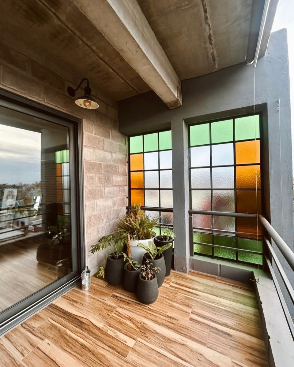 Balcón cubierto con tarima de madera, ventanas de vidrio esmerilado, plantas en macetas y puerta corrediza de vidrio.