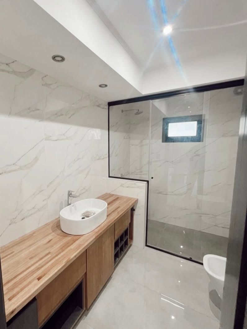 Baño moderno con tocador de madera, lavabo sobre encimera, ducha de cristal, paredes de mármol blanco