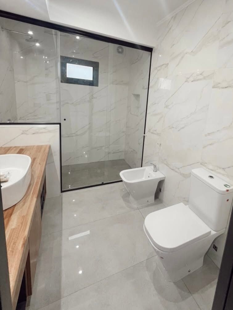 Baño moderno con ducha de cristal, bidé mural, inodoro y azulejos que imitan el mármol.