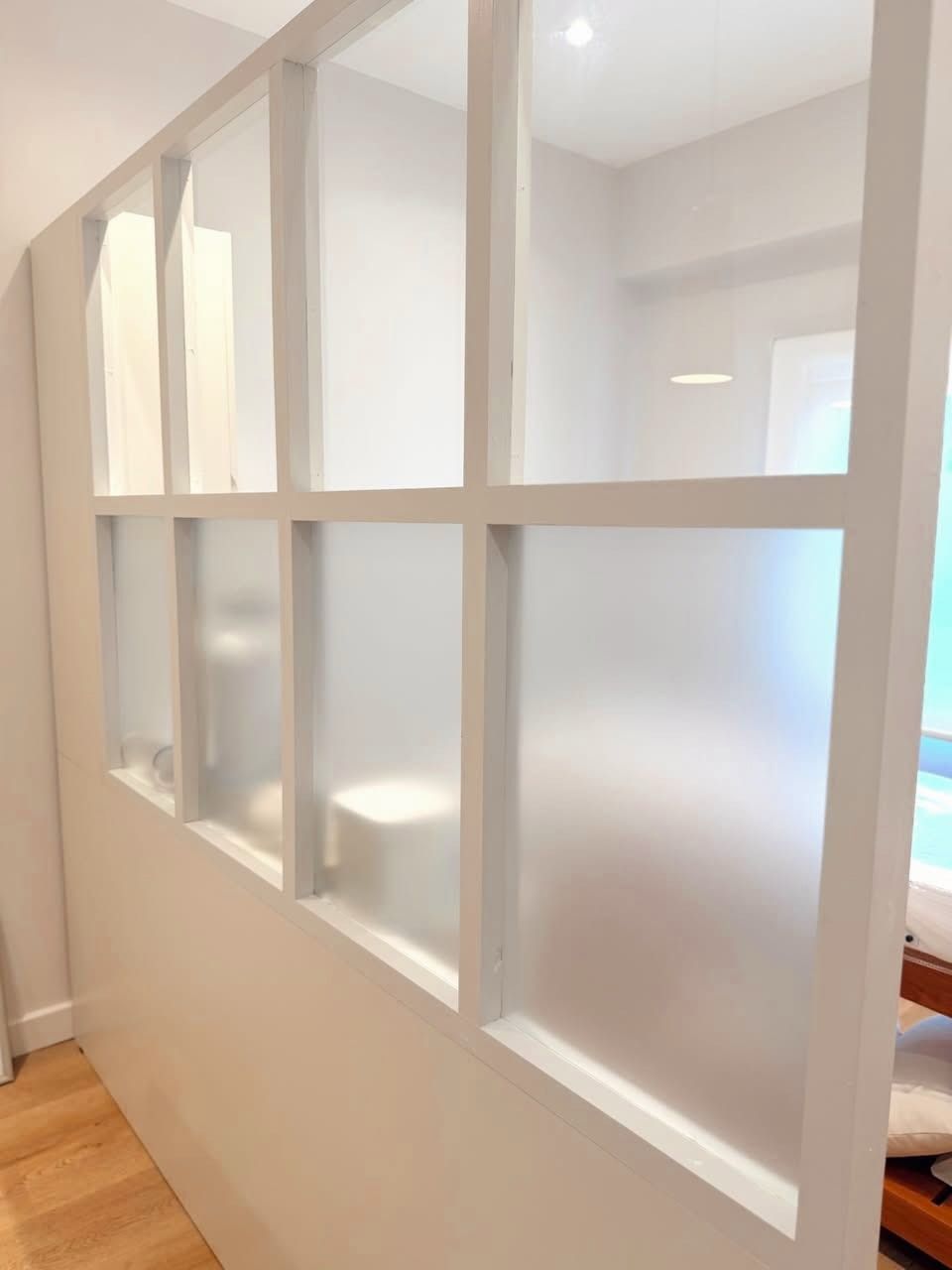 Pared interior con marco blanco y múltiples ventanas rectangulares recortadas, vista desde un pasillo con suelo de madera.