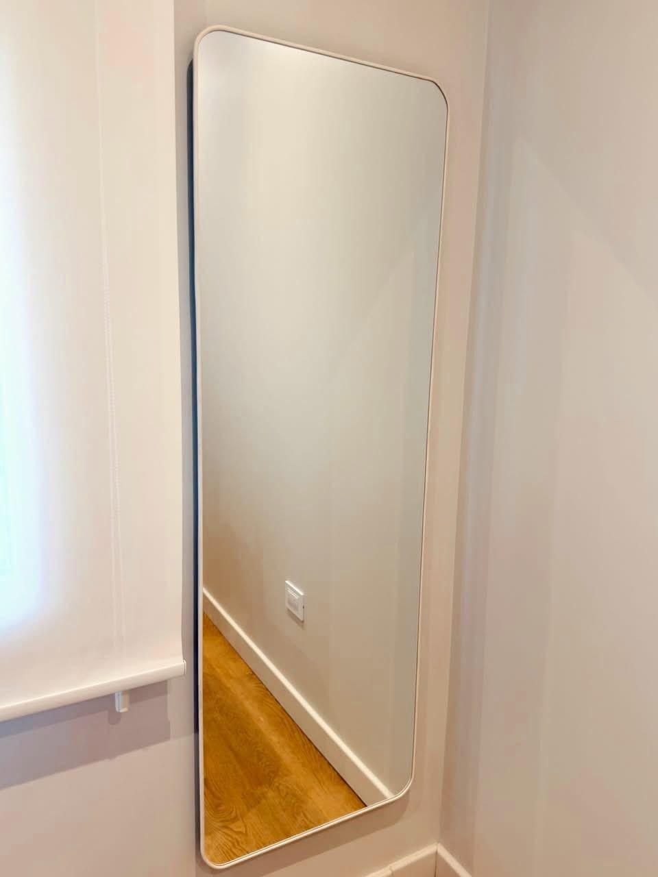 Un gran espejo de pared apoyado contra una pared blanca junto a una ventana, que refleja un suelo de madera.