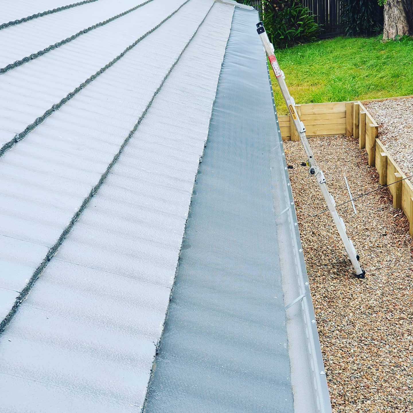 Metal roof beside a gravel path and wooden retaining wall, with green grass and a gutter downspout