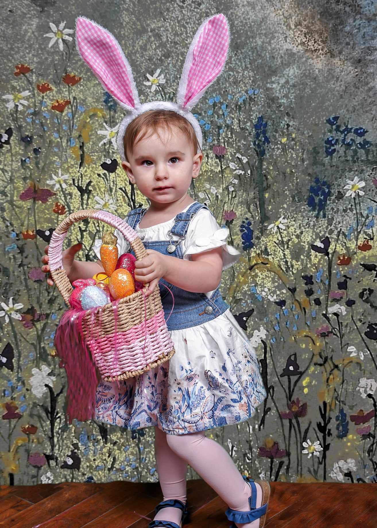 Child in bunny ears holding an Easter basket with colorful eggs in a floral garden backdrop