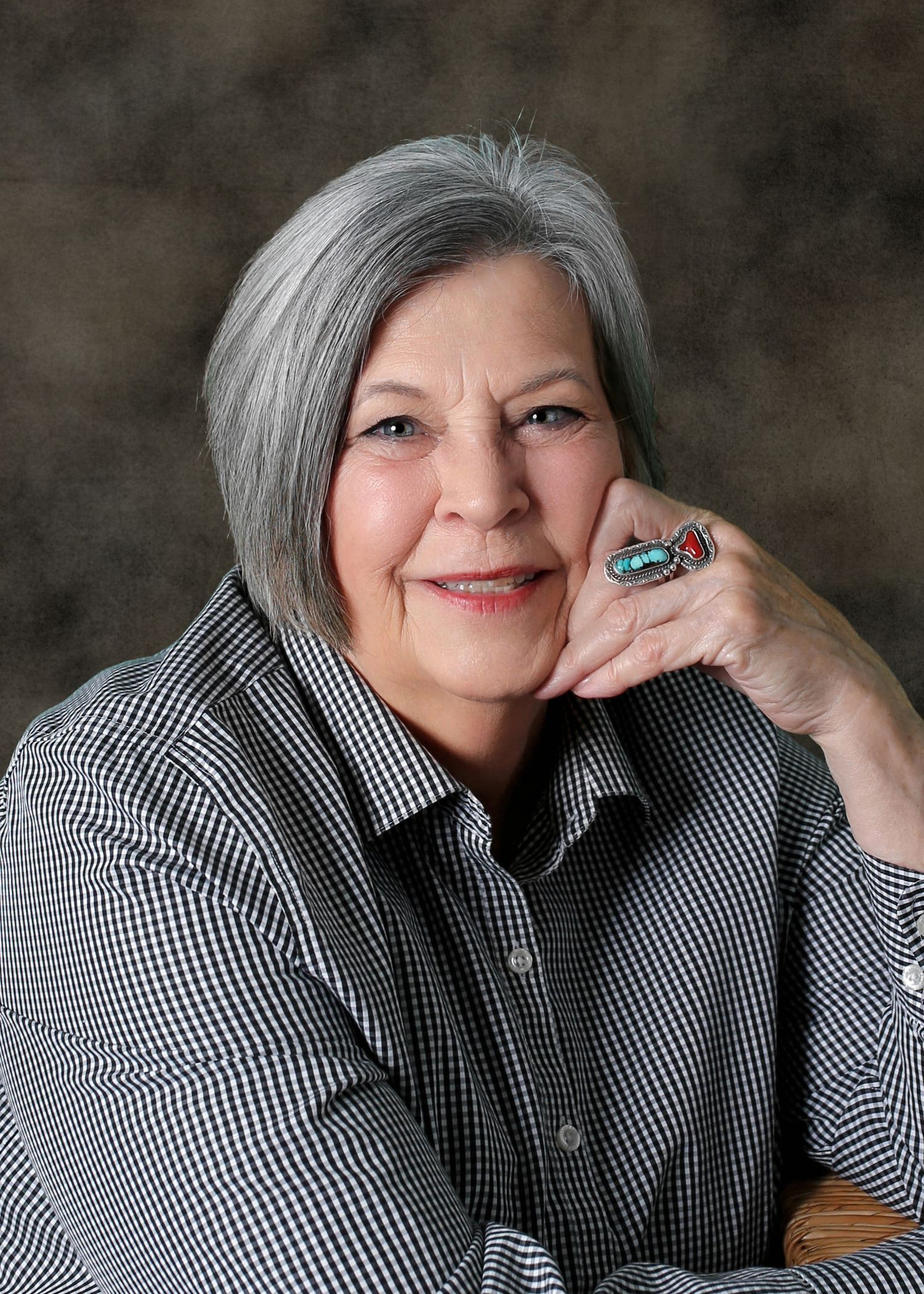 Portrait of a woman in a patterned shirt, smiling with her hand resting on her chin against a gray backdrop