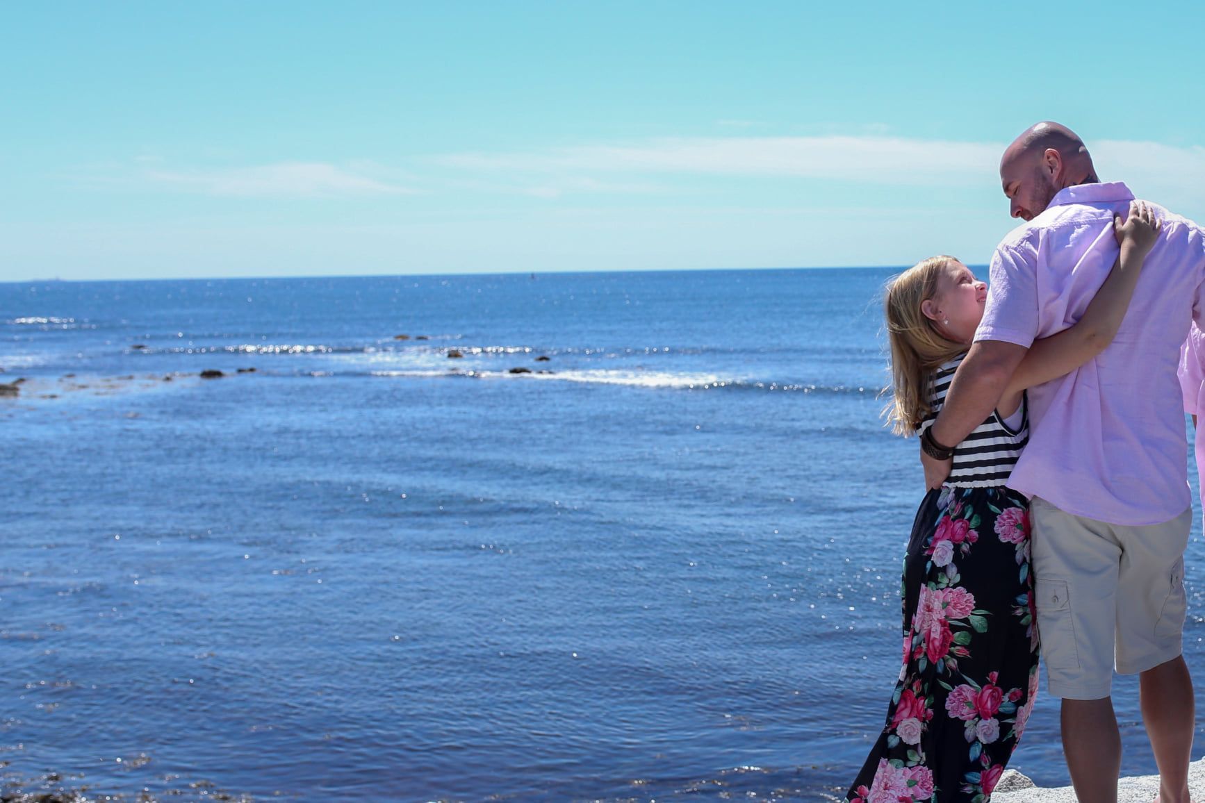 Couple embracing by the ocean on a sunny day, with blue water and a clear sky.