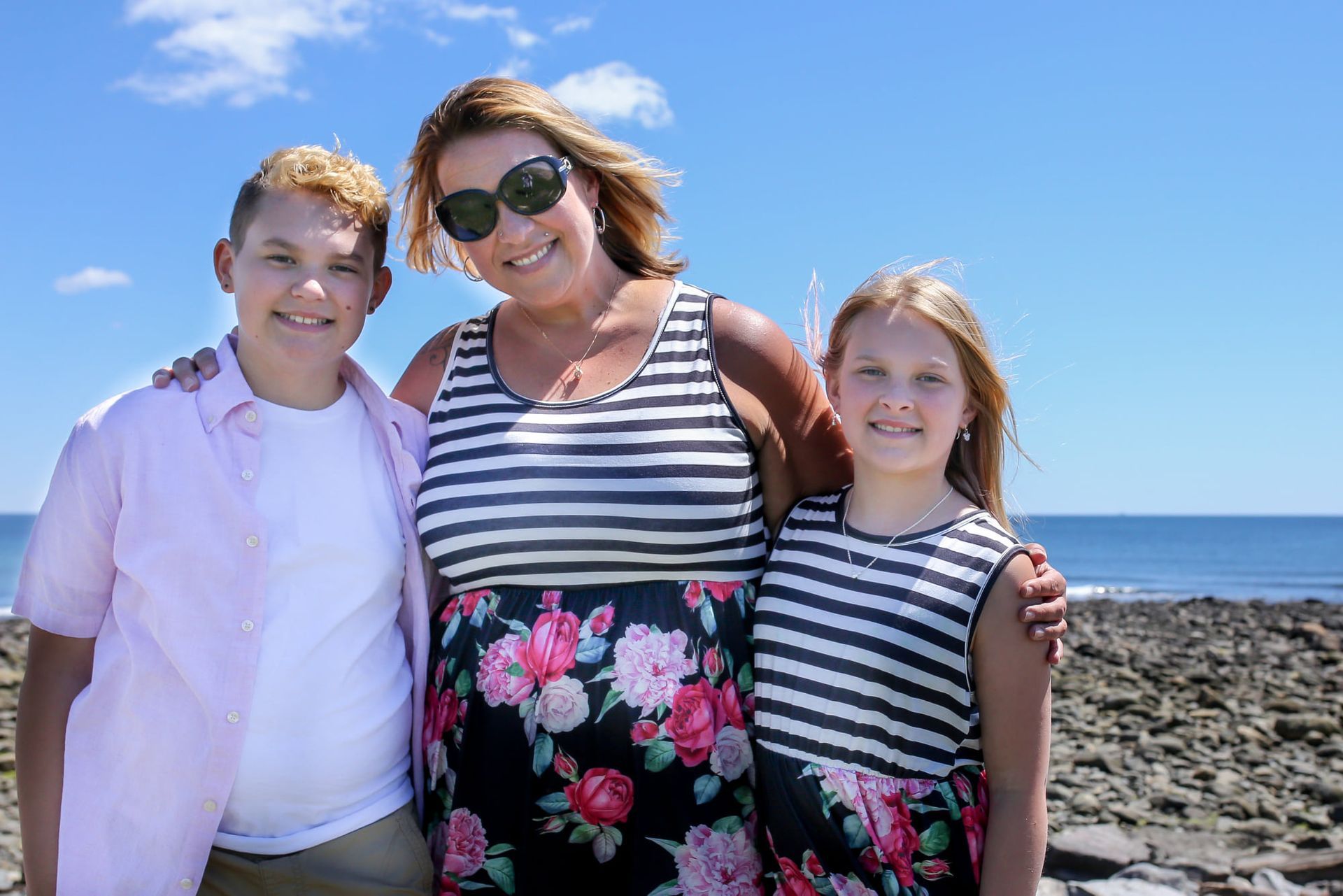 Three people posing on a rocky beach, smiling under a bright blue sky.